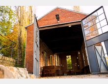 Backsteinbau mit großen geöffneten Metalltüren, die den Blick in einen renovierten Innenraum mit sichtbaren Ziegelwänden, hohen Fenstern und Holztheken freigeben. Im Vordergrund Herbstlaub und seitlich Bäume mit gelbem Laub.