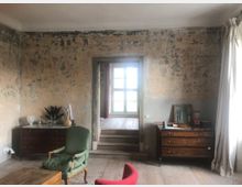 Ein Raum mit abgenutzten, verputzten Wänden, einem hellen Holzfußboden und einem großen Fenster in der Mitte des Hintergrunds. Im Vordergrund stehen zwei Kommoden aus dunklem Holz, ein grüner Polstersessel und ein Holztisch, während helle Vorhänge die rechte Seite des Bildes einrahmen.