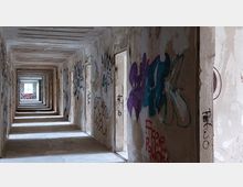 Langer, verfallener Korridor in einem verlassenen Gebäude, dessen Wände mit bunten Graffiti und Schriftzügen bedeckt sind. Der Korridor endet an einer stark beleuchteten Öffnung, durch die Tageslicht einfällt.