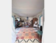 Großer Wohnraum mit bemustertem Fliesenboden und Teppichläufer, weißer Decke und hellen Wänden mit sichtbaren Gebrauchsspuren. In der Mitte stehen ein Couchtisch und Sitzmöbel; entlang der Wände befinden sich Schränke, Kommoden und Dekoration, flankiert von zwei blauen Fensterrahmen.