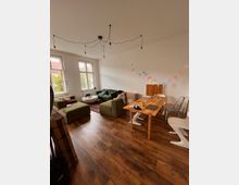 Ein Wohnzimmer mit Holzboden, einer Sitzgruppe aus grünen Sesseln und einem weißen Tischchen, sowie einem Holztisch mit verschiedenen Stühlen. An der Wand hängt eine farbige Wimpelkette, und die Deckenbeleuchtung besteht aus frei hängenden Glühbirnen. Tageslicht fällt durch hohe Fenster ein.