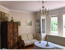 Wohnzimmer mit klassischer Einrichtung, darunter ein großer dunkler Holzschrank mit Verzierungen, ein weiß gestrichenes Vitrinenschrank an der Ecke, eine gepolsterte Sitzgarnitur und ein runder Tisch mit Tischdecke und zwei silbernen Kerzenhaltern. Große Fenster lassen viel Tageslicht herein, und an den Wänden hängen mehrere Bilder.