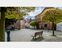 Ein kleiner, gepflasterter Platz in einer Altstadt, umgeben von historischen Backstein- und Putzgebäuden. Im Vordergrund stehen eine Holzbank, ein Brunnen und einige Bäume, während im Hintergrund Kirchenglocken als Dekoration auf dem Boden liegen.
