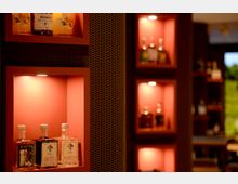 Innenansicht einer Bar mit in die Wand eingelassenen, warm beleuchteten Regalnischen, in denen mehrere Spirituosenflaschen ausgestellt sind. Zwischen den Nischen befindet sich eine dunkle Wandverkleidung mit metallischem Gittermuster, weitere Regale sind unscharf im Hintergrund zu sehen.