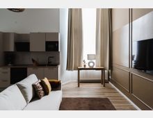 Wohnraum mit moderner Küchenzeile in neutralen Farben, einem grauen Sofa mit dekorativen Kissen, einem Holztisch vor einem großen Fenster mit beigefarbenen Vorhängen sowie einem Teppich auf einem Holzboden. An der Wand ist ein Flachbildfernseher angebracht.