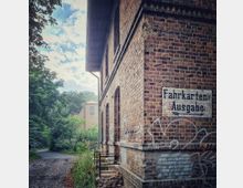 Backsteinbau mit der Aufschrift „Fahrkarten-Ausgabe“ an der Fassade, daneben ein schmaler, mit Kopfsteinpflaster belegter Weg, umgeben von Bäumen und Sträuchern. Im Hintergrund ragt ein weiteres Backsteingebäude hervor.