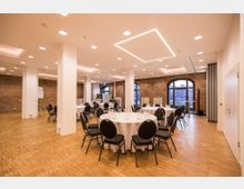 Ein moderner Veranstaltungsraum mit Backsteinwänden, großen Rundbogenfenstern und hellem Parkettboden. Rundtische mit weißen Tischdecken und gedecktem Geschirr sind gleichmäßig im Raum verteilt.