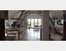 Ein geräumiges Wohnzimmer mit offenem Dachstuhl und sichtbaren Holzbalken. Der Raum enthält einen Esstisch mit Stühlen, eine Sitzgruppe in der Nähe eines großen Fensters, durch das ein Garten oder eine Landschaft zu sehen ist, sowie Schränke und Dekorationen aus Holz.