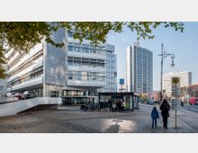 Straßenansicht in einer Stadt mit modernen Gebäuden, einer U-Bahnstation mit dem Schild „U-Bhf Ernst-Reuter-Platz“ im Vordergrund und mehreren Fahrradständern. Im Hintergrund stehen ein hohes Gebäude der Technischen Universität und weitere Hochhäuser, während links ein moderner Bürokomplex zu sehen ist.