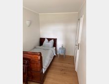 Schlafzimmer mit einem Einzelbett aus dunklem Holz, das an einer hellen Wand steht. Daneben ein kleiner, hellblauer Nachttisch mit einer Glaslampe; der Raum hat einen Holzboden und eine weiße Tür.