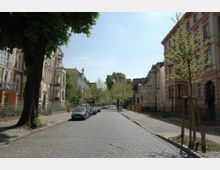 Kopfsteinpflasterstraße in einem Wohngebiet mit mehrstöckigen Altbauten auf beiden Seiten, teilweise mit verzierten Fassaden. Bäume säumen den Gehweg, und parkende Autos stehen entlang der Straße.