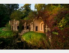 Steinbau mit Rundbögen und einer mit dichtem Efeu und roten Blättern bewachsenen Fassade, umgeben von grüner Vegetation und einem kleinen Teich im Vordergrund. Im Hintergrund sind Bäume erkennbar, die die Szene einrahmen.