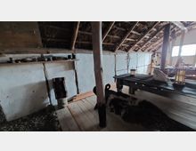 Innenraum eines historischen Fachwerkhauses mit sichtbaren Holzstützen und weiß verputzten Wänden. Zu sehen sind ein Holztisch mit Tierfellauflage und Kerzenlaternen, sowie ein Regal mit einfachen Gefäßen an der Wand.