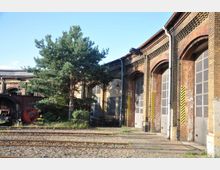 Backsteingebäude eines historischen Lokschuppens mit großen Toren, gelben Warnmarkierungen und angrenzenden Gleisen. Im Vordergrund steht ein Baum, und daneben ist ein Teil eines alten Eisenbahnrads zu sehen.