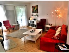 Wohnzimmer mit hellem Holzboden, einer roten Couch mit Kissen, einem passenden roten Sessel, einem weißen Couchtisch und einem roten Lesesessel neben einem kleinen Beistelltisch. Ein modernes Regal und eine Stehlampe befinden sich an der rechten Wand, während bodentiefe Fenster mit weißen Vorhängen den Raum erhellen.