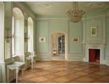 Prunkvoller Raum mit pastellgrünen Wänden, dekorativen Stuckelementen und Parkettboden. Auffällig sind ein Kronleuchter, ein dekorativer Kamin, wandhohe Spiegel sowie ein Durchgang mit Blick auf ein Geschirrschrankregal.