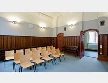 Ein kleiner Versammlungsraum mit hölzernen Wandverkleidungen, hellem Teppichboden und mehreren Holzstühlen in Reihen angeordnet. An den Wänden befinden sich Wandlampen, links eine Uhr, und rechts eine offene Tür, die zu einem benachbarten Raum mit Fenstern führt.