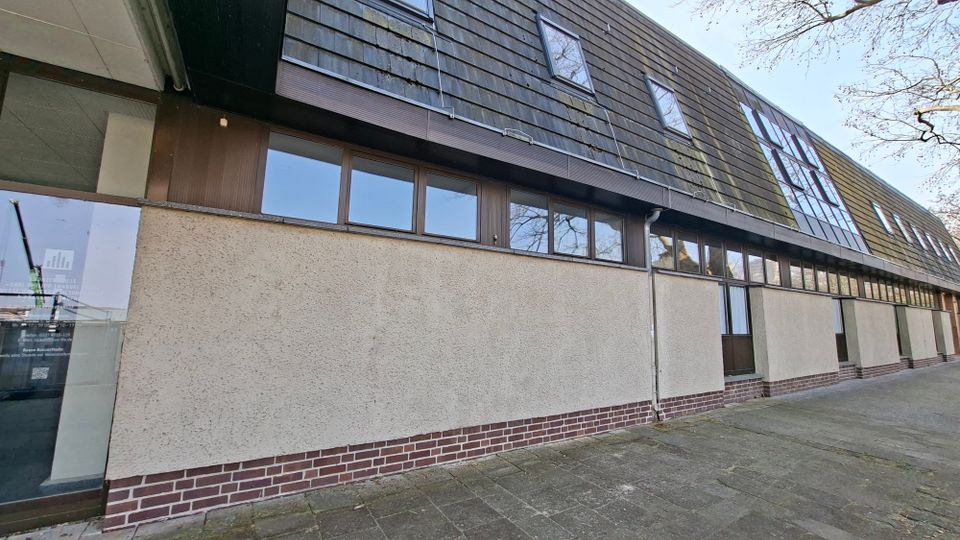 Außenansicht eines langgestreckten Gebäudes mit heller Putzfassade und Ziegelsockel, darüber eine dunkle, schieferartige Verkleidung mit Dachfenstern. Entlang der Fassade verläuft ein Band aus großen Fensterflächen und Türen; davor liegt ein gepflasterter Gehweg.