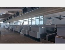 Leerer Check-in-Bereich in einem Flughafen mit grauen Schaltern und gelben Nummernschildern (B34-B36). Im Hintergrund sind große Fenster mit diffusem Tageslicht sowie eine Überdachung mit gitterartiger Verkleidung zu sehen.