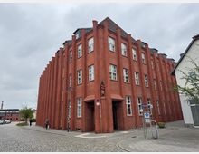 Rotes Backsteingebäude mit modernen Stilelementen, hohen vertikalen Fassadenvorsprüngen und mehreren symmetrisch angeordneten weißen Fenstern, an einer gepflasterten Straßenecke. Im Vordergrund befinden sich Fahrradständer, Straßenschilder und eine Gehwegfläche.