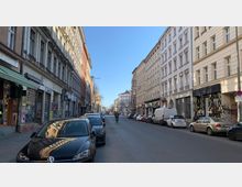 Straßenszene in einer Stadt mit mehrstöckigen historischen Wohn- und Geschäftsgebäuden auf beiden Seiten. Geparkte Autos und ein Radfahrer befinden sich auf der asphaltieren Straße, die von Geschäften und Fassaden mit klassischer Architektur gesäumt ist.