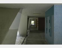 Überdachter Gang mit Betonpfeilern und Geländer, flankiert von blauen, gefliesten Wänden auf der rechten Seite und schrägen Betonflächen auf der linken Seite. Im Hintergrund ist ein heller Außenbereich sichtbar.