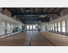 Großer, lichtdurchfluteter Saal mit Holzfußboden und industriellem Charme, geprägt von einer Betondecke, sichtbaren Rohren und einer Ziegelwand. An den großen Fenstern stehen Pflanzen, und in der rechten Ecke führt eine Wendeltreppe nach oben.