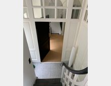 Treppenhaus in einem Innenraum mit hellen Wänden und Glaselementen in den Türen. Sicht auf eine geöffnete schwarze Tür, dahinter ein Raum mit Teppichboden sowie eine angrenzende Treppe mit einem schwarzen Handlauf.