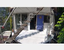 Ein Hausboot mit einer Metalltreppe an der Vorderseite, die zu einer oberen Ebene führt. Davor befinden sich eine blaue Tür mit Bullauge, vergitterte Fenster und Pflanzen im Hintergrund; das Boot ist an einem Flussufer gelegen.