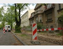 Straßenszene mit Baustelle in einer Wohngegend; links verläuft eine gepflasterte Straße mit geparktem Kleintransporter, rechts stehen ältere Häuser mit teilweise renovierungsbedürftigen Fassaden, während Absperrbaken und aufgerissener Gehweg auf Bauarbeiten hinweisen.