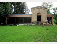 Backsteingebäude mit klassizistischer Fassade und angrenzendem Anbau aus Holz mit großen Glasfenstern in einer grünen Parklandschaft. Vor dem Gebäude stehen Gartenmöbel und Klappstühle auf einer Rasenfläche, umgeben von Bäumen.