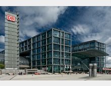 Moderne Glasfassade des Berliner Hauptbahnhofs mit mehreren Gebäudeteilen und einem weithin sichtbaren „DB“-Logo auf einem Turm. Vor dem Bahnhof befinden sich ein großzügiger Platz, Besucher und geparkte Fahrzeuge.