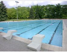 Ein großes, rechteckiges Freibad mit markierten Schwimmbahnen, umgeben von Gehwegen und Bäumen im Hintergrund. Am Beckenrand stehen einzelne Startblöcke, und das Wasser reflektiert den Himmel.