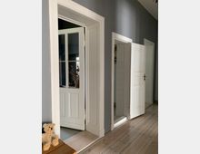 Ein Flur in einem Wohnhaus mit grauen Wänden und Holzboden; links steht eine offene Glastür mit weißem Rahmen, rechts daneben zwei weiße Holztüren, von denen eine geöffnet ist. Auf einer Holzkommode links liegt ein Stofftier.