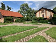 Zwei historische Backsteingebäude in einer ländlichen Umgebung, ein Satteldach-Stallgebäude mit roten Ziegeln links und ein größeres, verputztes Haus rechts. Im Vordergrund befinden sich grüne Rasenflächen und gepflasterte Wege, umgeben von Bäumen und Sträuchern.
