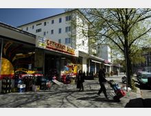 Straßenszene mit einem Getränkemarkt, erkennbar an der Beschriftung "Getränke Soley", und Getränkekisten vor dem Eingang. Im Vordergrund ein Fußgänger mit einer Sackkarre, im Hintergrund ein Wohngebäude und geparkte Autos entlang der Straße, gesäumt von Bäumen mit grünem Laub.