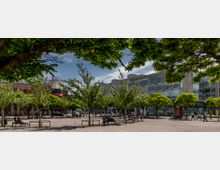 Ein öffentlicher Platz mit Bäumen und Sitzbänken im Vordergrund, umgeben von modernen Gebäuden mit Glas- und Steinfassaden. Menschen sitzen, spazieren oder fahren Fahrrad, während der Platz von schattenspendendem Grün eingerahmt wird.