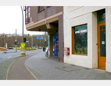 Straßenszene mit Gehweg, einer Häuserfassade und Schaufenster eines Geschäfts mit der Aufschrift „Afrikanische Afro Kunst“. Im Hintergrund Verkehrsampeln, eine Kreuzung und Straßenschilder, die Richtung „Schöneberg“ weisen.