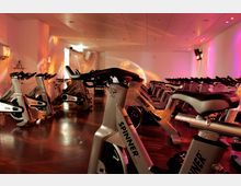 Ein Fitnessraum mit mehreren stationären Fahrrädern (Spinning-Bikes) in Reihen angeordnet. Der Raum hat eine warme Beleuchtung mit farbigen Lichtprojektionen in Rosa und Orange auf weißen Wänden und einem glänzenden Holzboden.