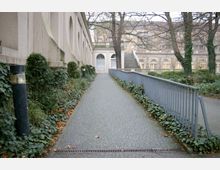 Ein gepflasterter Weg führt entlang einer beigefarbenen Fassade mit Rundbögen, umgeben von Efeu und kleinen Sträuchern. Rechts des Weges befindet sich ein Metallgeländer, und im Hintergrund sind Fenster eines Gebäudes sowie eine Treppe zu sehen.