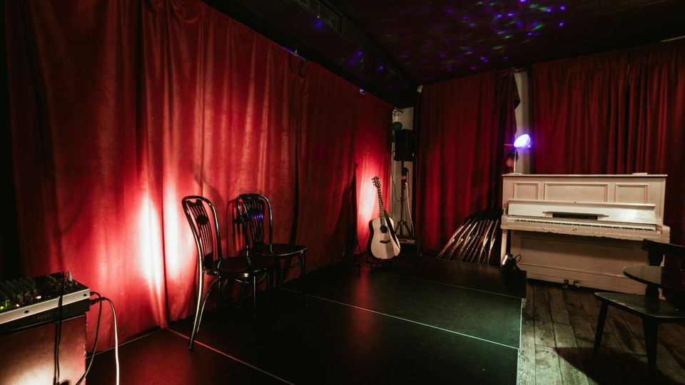 Small performance room with a low stage in front of red velvet curtains, lit by colored spotlights on a dark ceiling. A white upright piano, an acoustic guitar on a stand, stacked chairs, and audio equipment sit along the sides on a wooden floor.