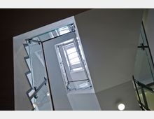 Blick nach oben in einem modernen Treppenhaus mit gläsernen Geländern und Metallrahmen; durch das spitzwinklige Treppenhaus ist ein rechteckiges, verglastes Oberlicht zu sehen, das Tageslicht hereinscheint.