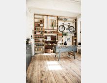 Industrieller Raum mit Holzboden, Regalen gefüllt mit Büchern, Flaschen und Dekorationsgegenständen, darunter ein Fahrrad und eine alte Glocke. Im Vordergrund steht ein Tischkicker, und die Wände sind aus weiß gestrichenem Backstein.