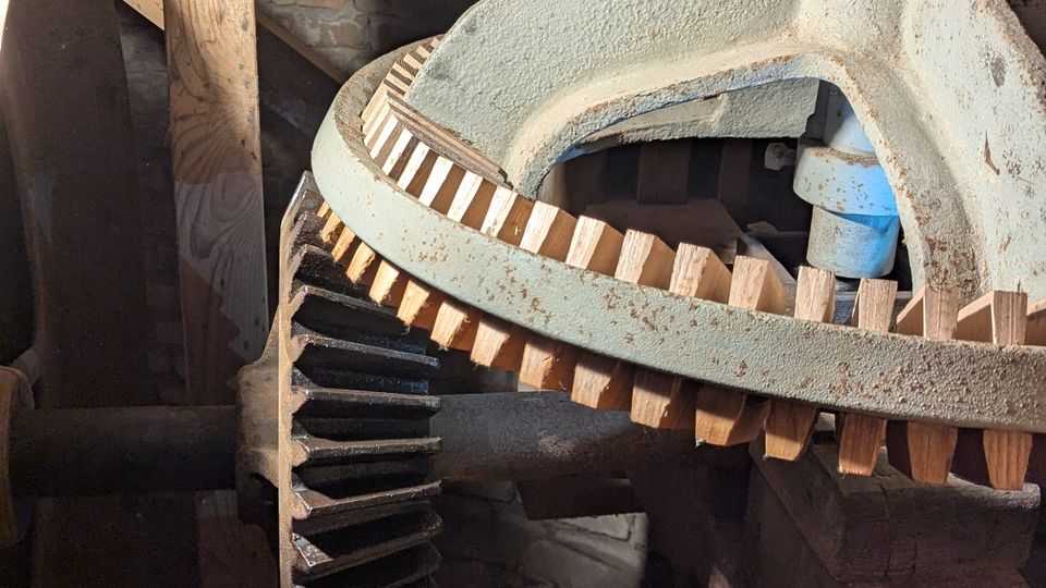 Blick in den Innenraum eines alten Holzgebäudes mit dicken Holzbalken und einer gemauerten Wand im Hintergrund. Im Vordergrund ist ein großes, teilweise verrostetes Zahnradgetriebe aus Metall mit Holz-Zahnkranz und einer senkrechten Welle zu sehen.