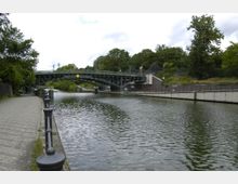 Fluss mit Uferpromenade und schmiedeeiserner Brücke, umgeben von Bäumen und Grünflächen. Die Promenade ist mit Kopfsteinpflaster versehen, mit schwarz lackierten Geländern abgegrenzt, während die Brücke moderne Geländerelemente und Gelbleuchten zeigt.