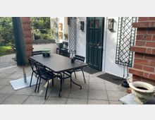 Außenbereich mit einem gepflasterten Boden, einem schwarzen Tisch und mehreren Stühlen. Im Hintergrund ist eine weiße Hausfassade mit grüner Tür, Rankgittern und Fenstern zu sehen, daneben befinden sich Ziegelwände und ein bepflanzter Gartenbereich.