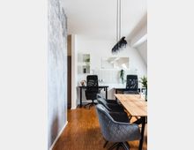 Modern eingerichteter Arbeits- und Essbereich mit Holzfußboden, einem Esstisch aus Holz, umgeben von grauen Polsterstühlen, und zwei Schreibtischen mit schwarzen Bürostühlen im Hintergrund. An der Wand sind ein Spiegel, kleine Regale und eine Lochplatte mit Dekorationen platziert. Über dem Esstisch hängt eine moderne Hängeleuchte mit mehreren schwarzen Glaskugeln.