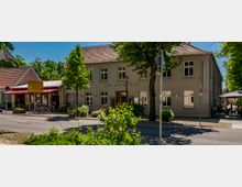 Straßenszene mit einem zweigeschossigen, hell verputzten Gasthaus mit Satteldach und der Aufschrift „Zum Alten Fritz“, umgeben von Bäumen. Vor dem Gebäude stehen Fahrräder und Straßenschilder, rechts ist eine Außengastronomie mit Tischen und Sonnenschirmen zu sehen.
