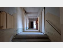 Langer, schmaler Innenflur in einem stark abgenutzten Gebäude mit abblätternder Farbe, niedriger Decke und kleinen Stufen im Vordergrund. Links hängt ein großer Heizkörper, rechts führt ein Treppengeländer an offenen Türen entlang zu einem helleren Bereich am Ende des Gangs.