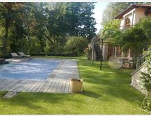 Gartenansicht mit einem überdachten Swimmingpool in der Mitte, umgeben von einer Rasenfläche, Liegestühlen und Bäumen. Rechts im Bild befindet sich ein Gebäude mit großen Fenstern, einer Außentreppe und einer Terrasse mit Gartenmöbeln.